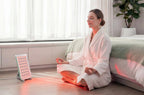 Woman in a white robe using nanoleaf red light therapy device on her leg in a bedroom.
